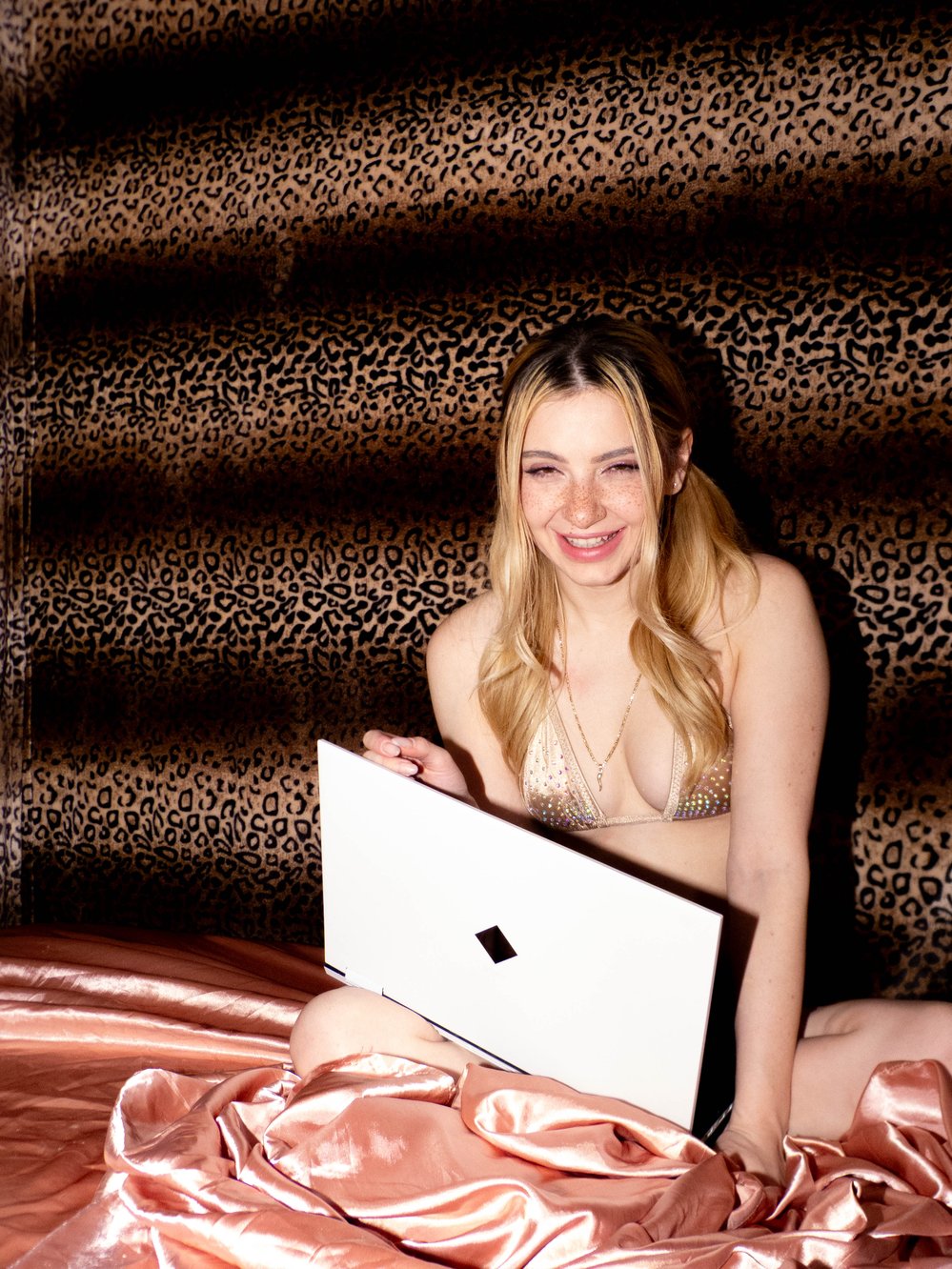 Demi Hawks sits with a laptop in a gold bikini top against a leopard-print backdrop.