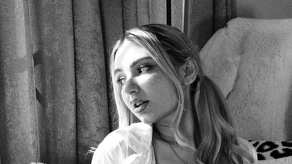 Demi Hawks looks off-camera in an open white shirt, freckles visible, black and white.