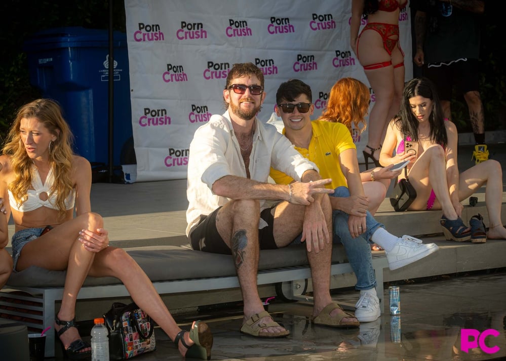Guests, with Girthmasterr in the middle, relax in front of the PornCrush step-and-repeat backdrop during Media Day 2026.