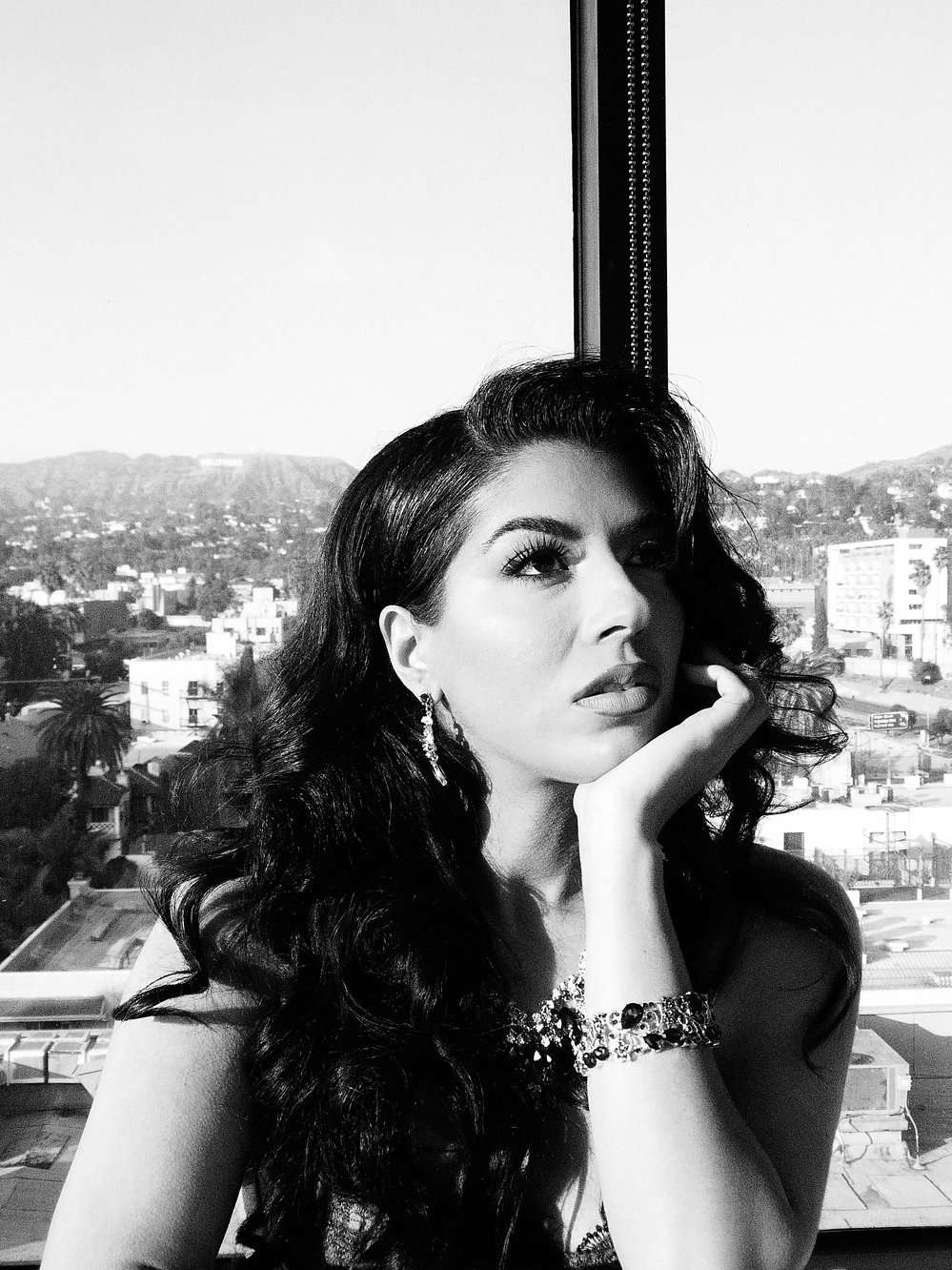 A black-and-white portrait of Jasmine Sherni resting her chin on her hand before a Los Angeles skyline window.